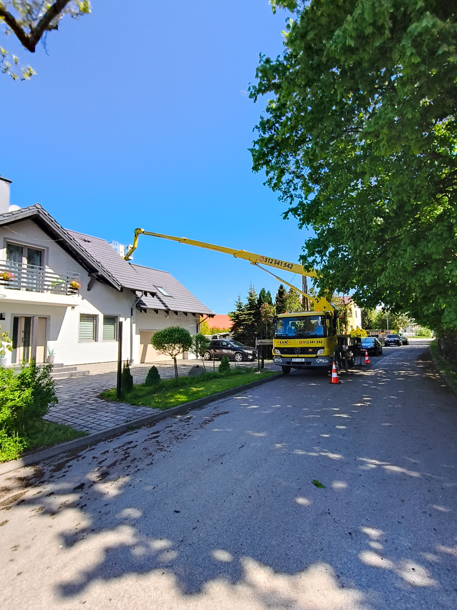 Żółty podnośnik koszowy przy białym domu z szarą dachówką, na tle błękitnego nieba. Widoczne drzewa i zaparkowane samochody. Ujęcie z poziomu ulicy.
