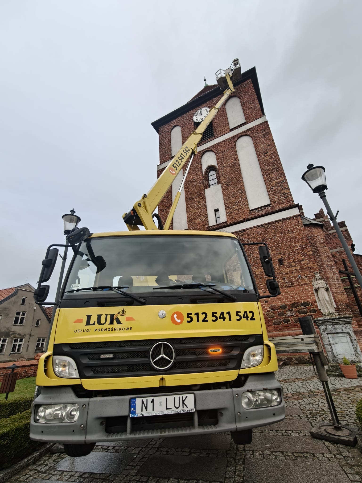 Żółty podnośnik koszowy LUK na tle ceglanej wieży kościoła z zegarem. Kosz podnośnika na wysokości zegara. Widoczny numer telefonu i logo firmy.
