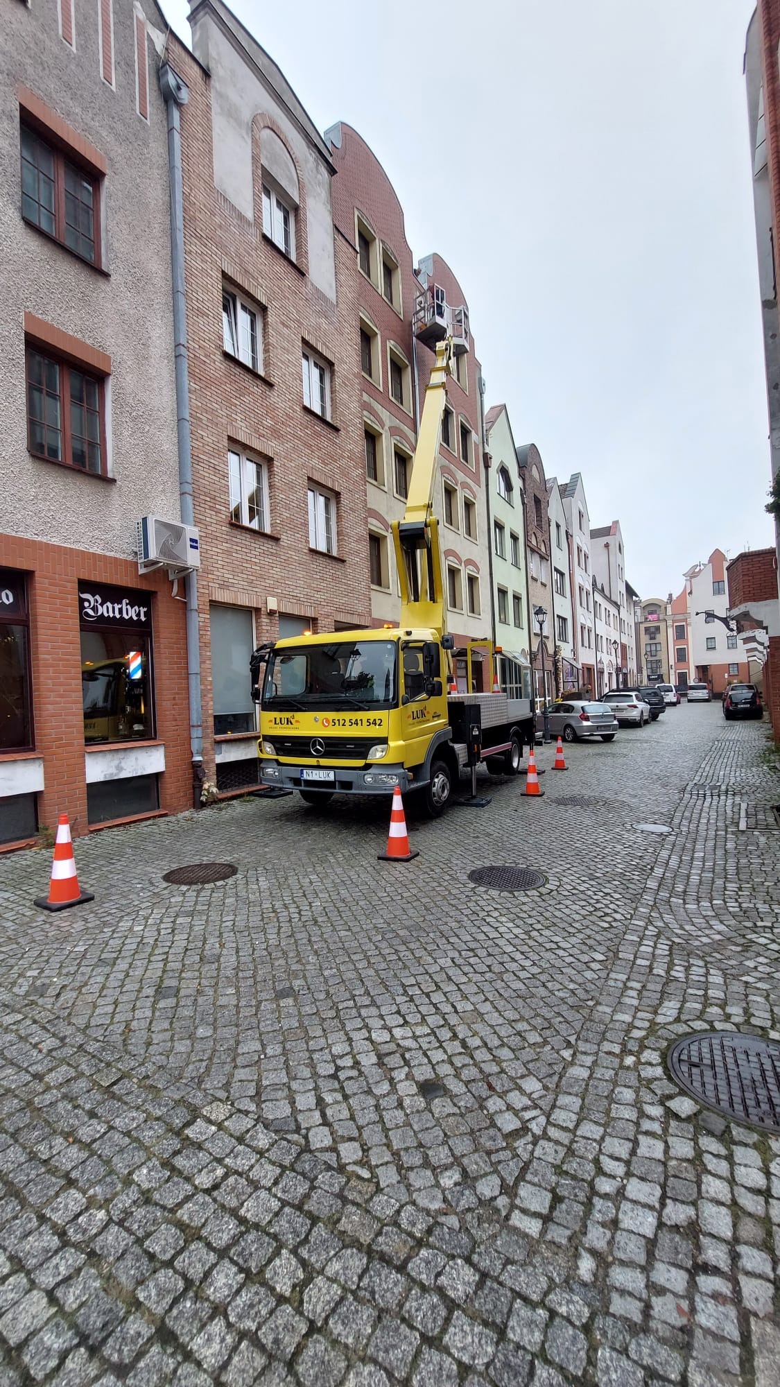 Żółty podnośnik koszowy LUK na brukowanej ulicy, z wysięgnikiem sięgającym do okna kamienicy. Pomarańczowe pachołki zabezpieczają obszar robót. Widok na starą zabudowę miejską.