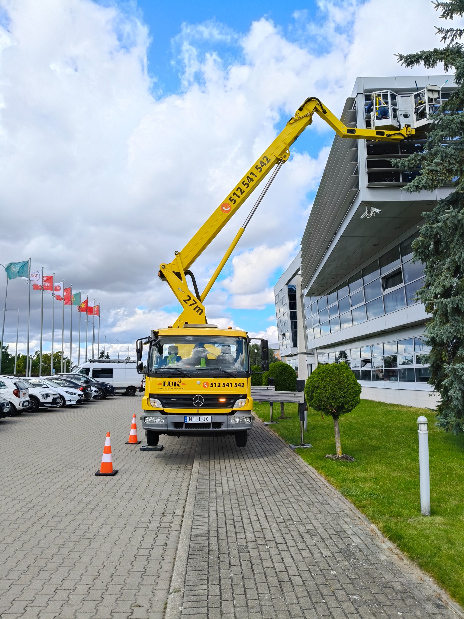 Żółty podnośnik koszowy LUK na chodniku, z pracownikami w koszu przy szklanej fasadzie budynku. Flagi w tle, oznakowanie stożkami.