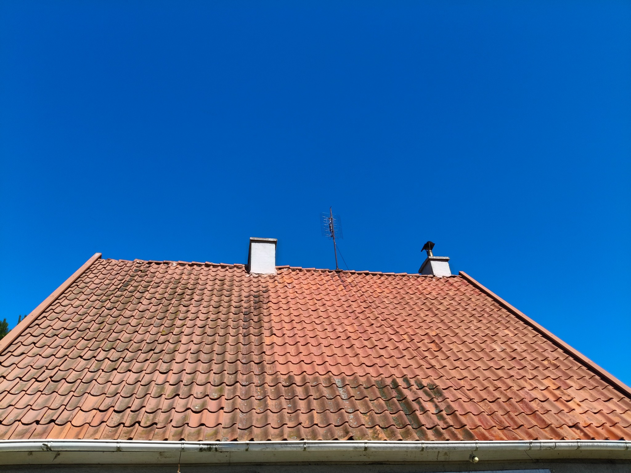 Czerwona dachówka z anteną i kominami na tle błękitnego nieba. Widoczne ślady zabrudzeń na dachu. Ujęcie z dołu, podkreślające strukturę i kolor dachu.