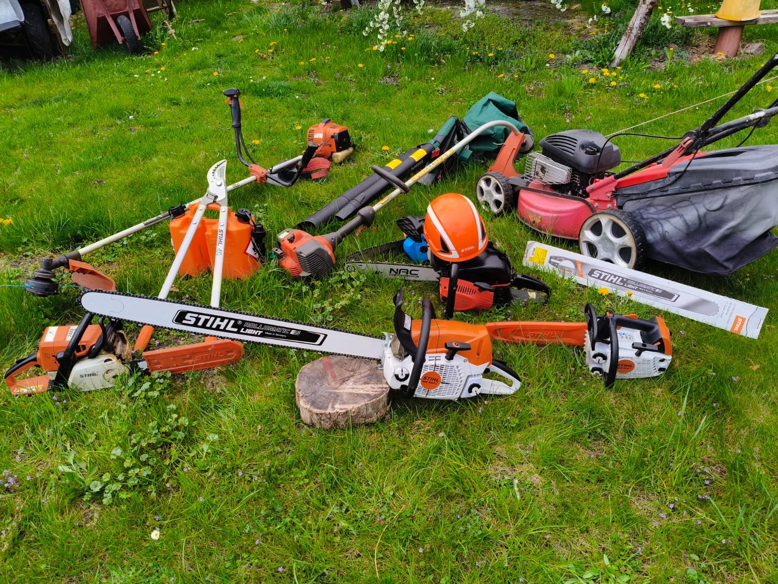 Zestaw narzędzi ogrodniczych Stihl na trawie: pilarki, kosiarki, nożyce, dmuchawa, kanister i kask. Sprzęt gotowy do pracy w ogrodzie, w tym pilarka oparta o drewniany pieniek.