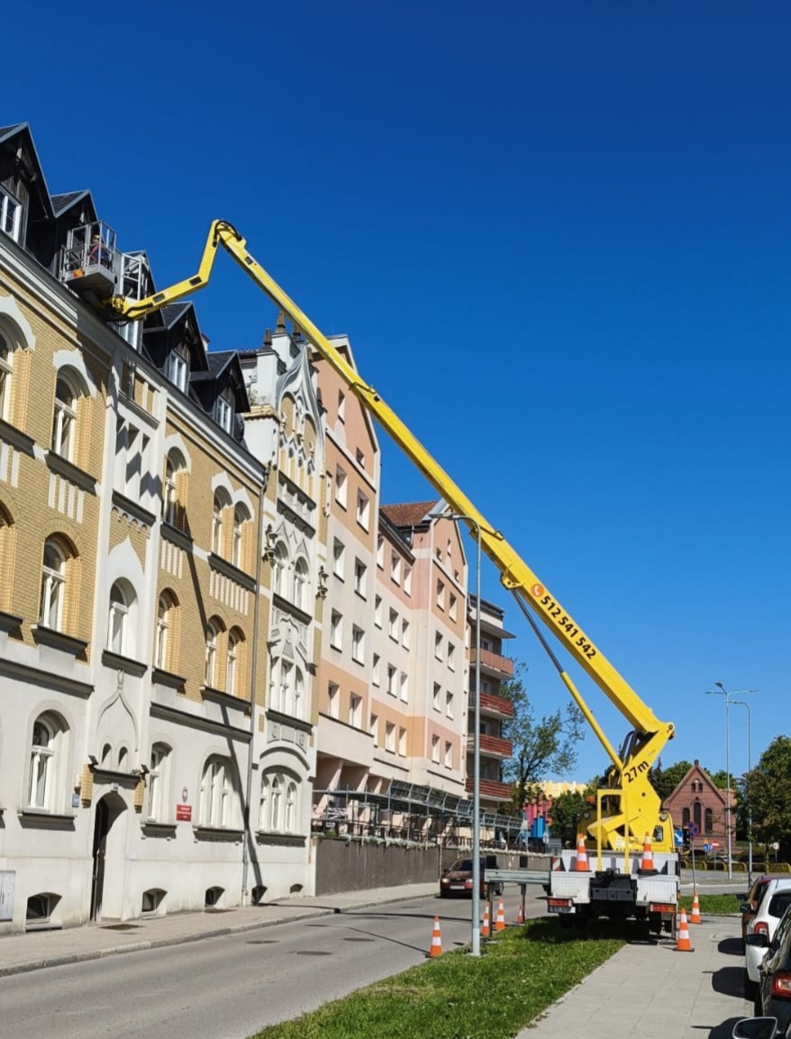Żółty podnośnik koszowy na tle kamienicy z osobą w koszu, błękitne niebo. Widoczny numer telefonu na ramieniu podnośnika.