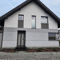 ZUNN - Dwukondygnacyjny dom z szarą elewacją i ciemnym dachem, widoczny z poziomu chodnika z kostki brukowej. Otwarta brama wejściowa. Minimalistyczny design.