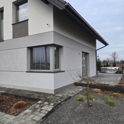 ZUNN - Nowoczesny dom z szarą elewacją i kostką brukową. Widoczny fragment ogrodu z kamieniami i małym drzewkiem. Ujęcie z dołu, podkreślające geometrię budynku.