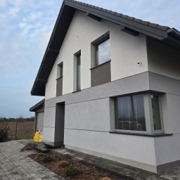 ZUNN - Nowoczesny dom jednorodzinny z elewacją w odcieniach szarości i białym wykończeniem. Widoczny fragment chodnika z kostki brukowej i niewielki ogródek.