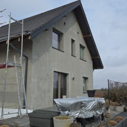 ZUNN - Dom w trakcie prac elewacyjnych: szare ściany, rusztowanie z boku. Widoczne okna i dach. Na pierwszym planie elementy ogrodu.