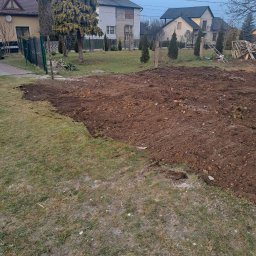 Olbud - Przygotowany teren pod budowę na działce z widokiem na domy jednorodzinne. Częściowo usunięta darń, odsłonięta brązowa ziemia. Widoczne ogrodzenie i drzewa.