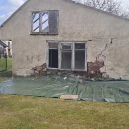 Olbud - Stary, zaniedbany budynek z uszkodzoną elewacją i oknami, zabezpieczony zieloną plandeką. Widoczne pęknięcia i ubytki w ścianie. Częściowo zabite okno na poddaszu.