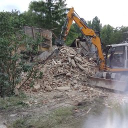 Olbud - Żółta koparka Liebherr z osprzętem do rozbiórki, aktywnie usuwająca gruz z częściowo zburzonego budynku, otoczonego zielenią pod zachmurzonym niebem.