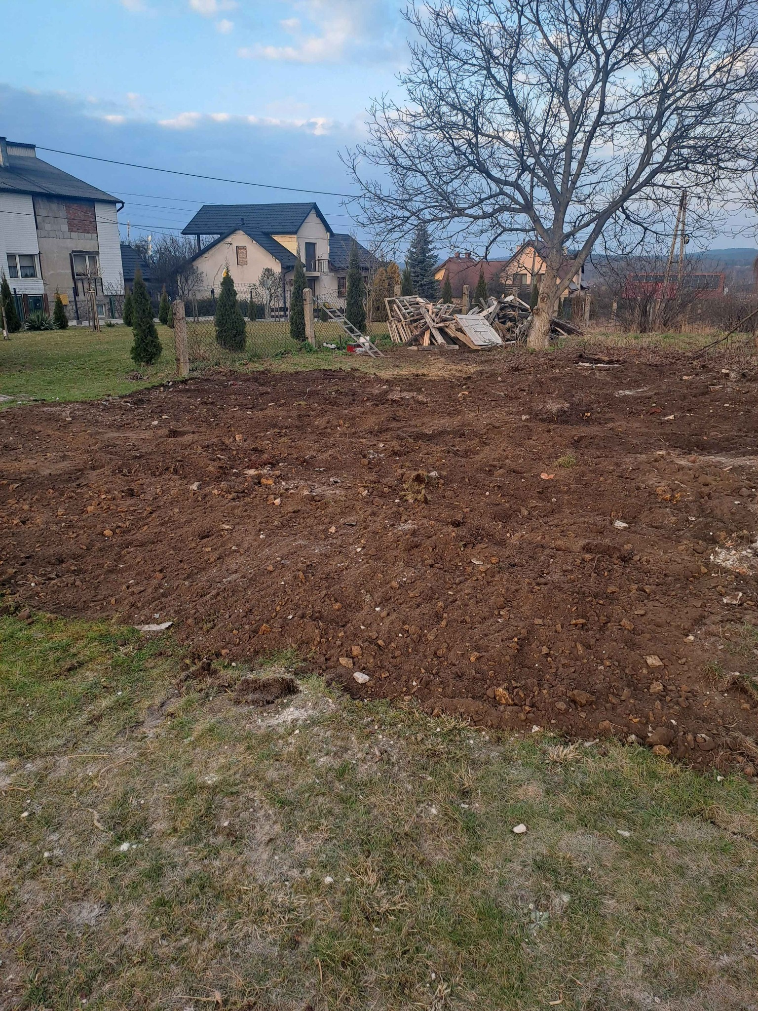 Przygotowany teren pod budowę domu jednorodzinnego. W tle widoczne domy, drzewa i pozostałości po rozbiórce. Ziemia wyrównana, gotowa pod fundamenty.