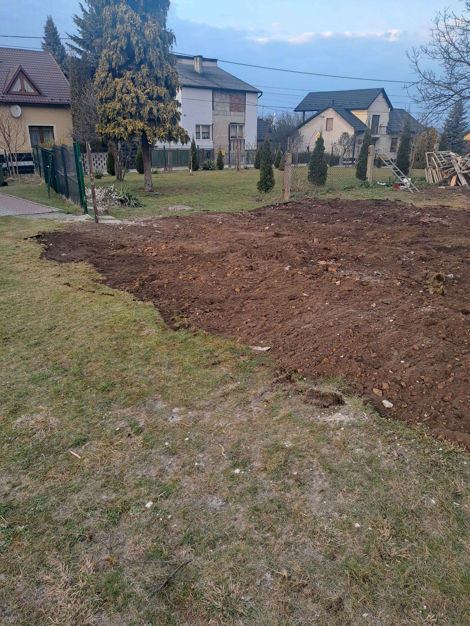 Przygotowany teren pod budowę na działce z widokiem na domy jednorodzinne. Częściowo usunięta darń, odsłonięta brązowa ziemia. Widoczne ogrodzenie i drzewa.