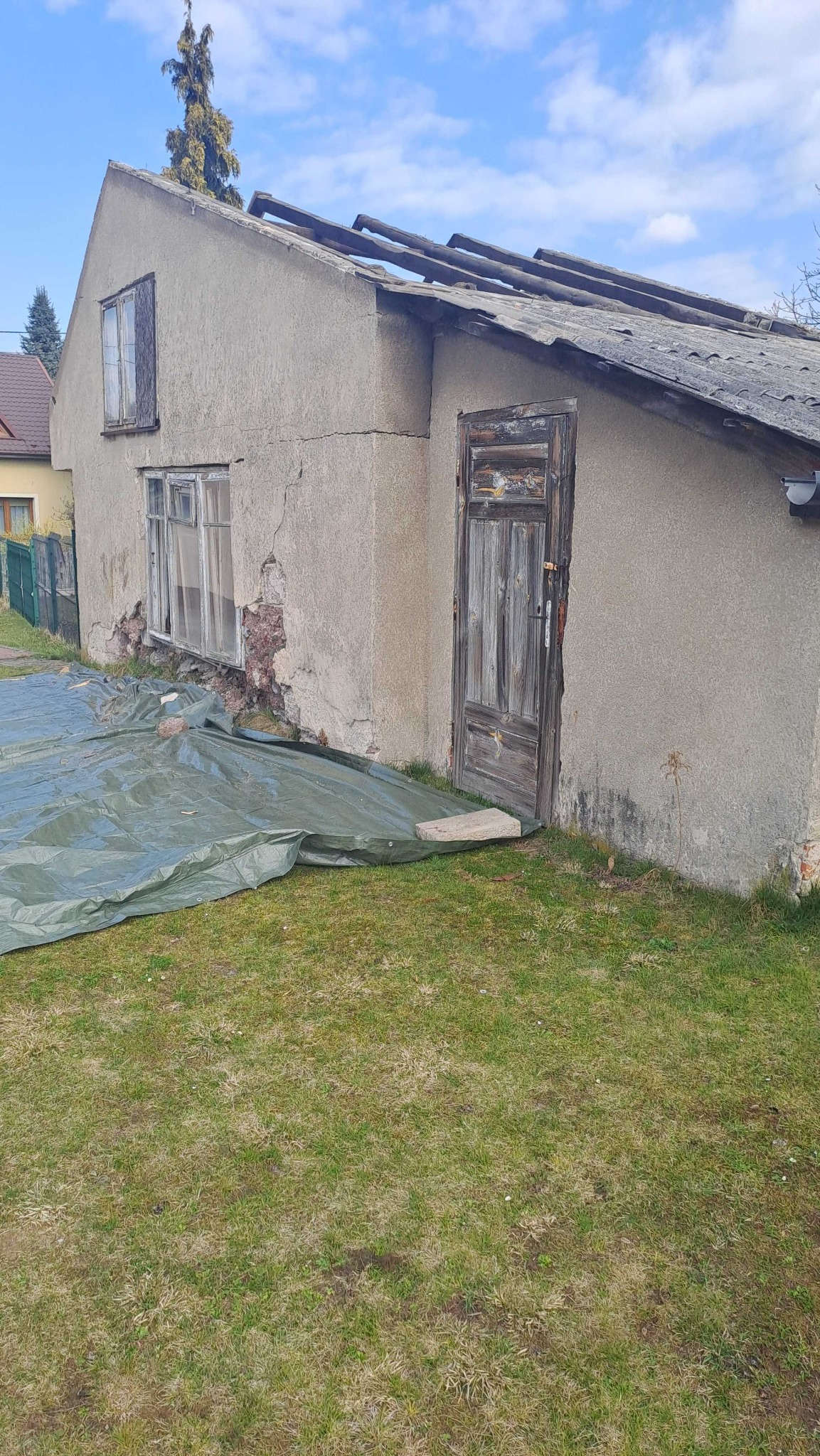 Stary, częściowo zdemontowany budynek z uszkodzoną elewacją i dachem, zabezpieczony plandeką. Widoczne odsłonięte elementy konstrukcyjne i zniszczone okna.