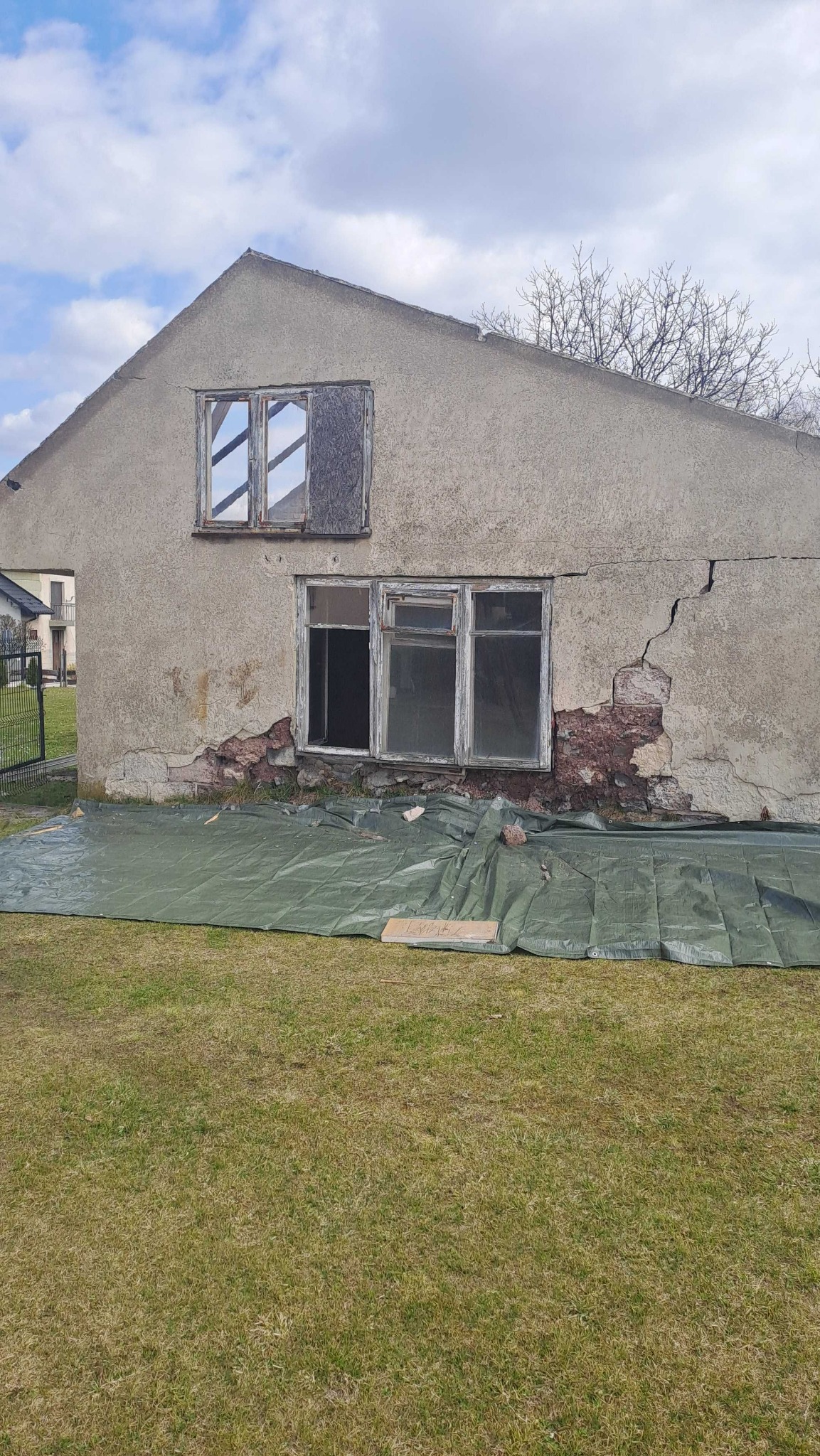 Stary, zaniedbany budynek z uszkodzoną elewacją i oknami, zabezpieczony zieloną plandeką. Widoczne pęknięcia i ubytki w ścianie. Częściowo zabite okno na poddaszu.