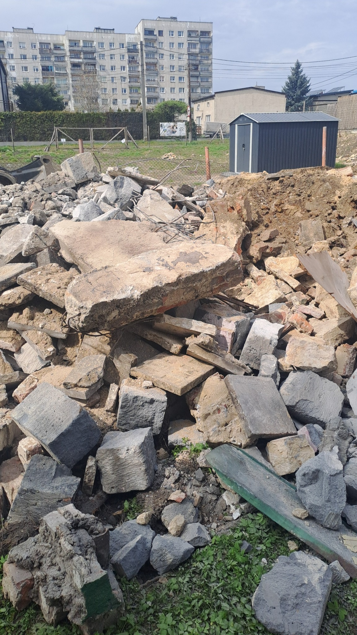 Gruzowisko z fragmentami betonu i cegieł na tle budynku mieszkalnego i blaszanej budki. Widoczne pozostałości po rozbiórce.