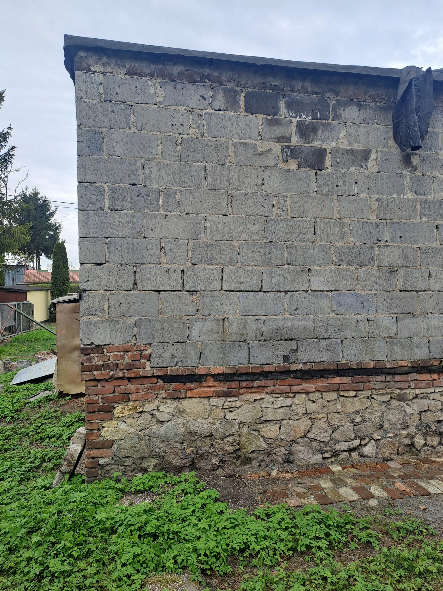 Fragment ściany budynku z szarej cegły, z widocznymi warstwami cegły czerwonej i kamiennej u podstawy. W tle zieleń i fragmenty zabudowań.