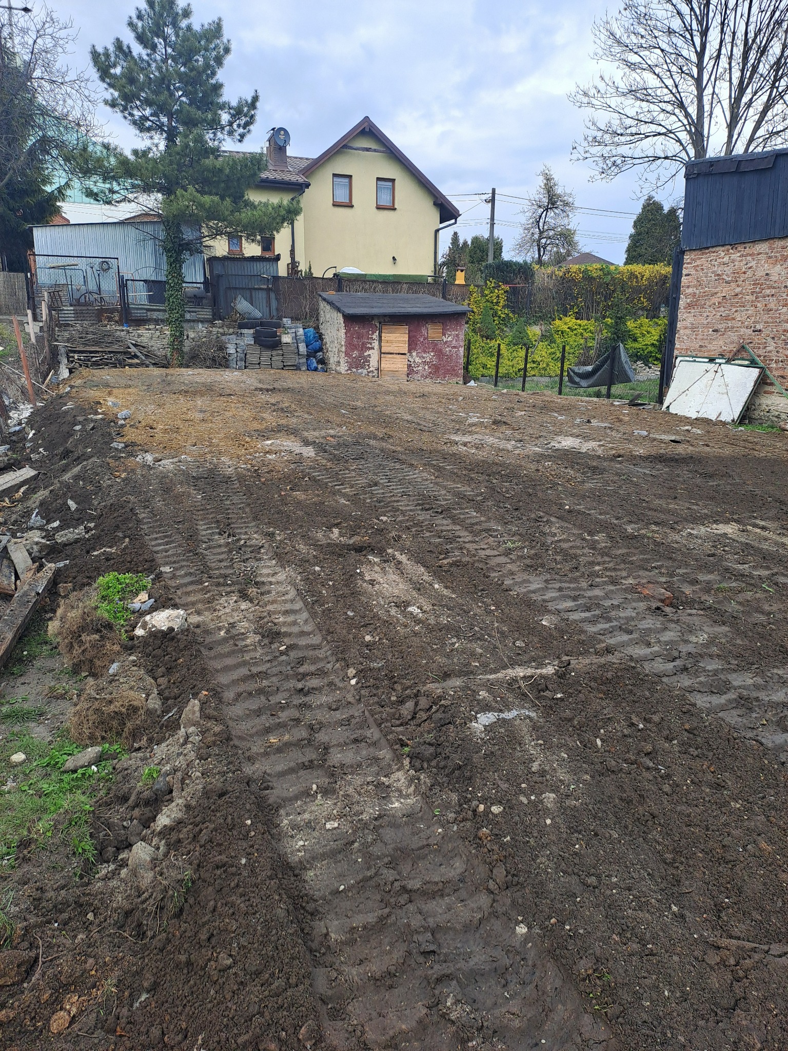 Przygotowany teren pod budowę z widocznymi śladami opon maszyny budowlanej. W tle zabudowania mieszkalne i gospodarcze. Częściowo usunięta warstwa ziemi.