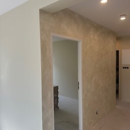 TopTynk TK - Beżowa ściana z efektem dekoracyjnym w nowym wnętrzu. Widoczne gniazdka elektryczne i fragment podłogi. W tle otwarte drzwi i ułożone kartony.