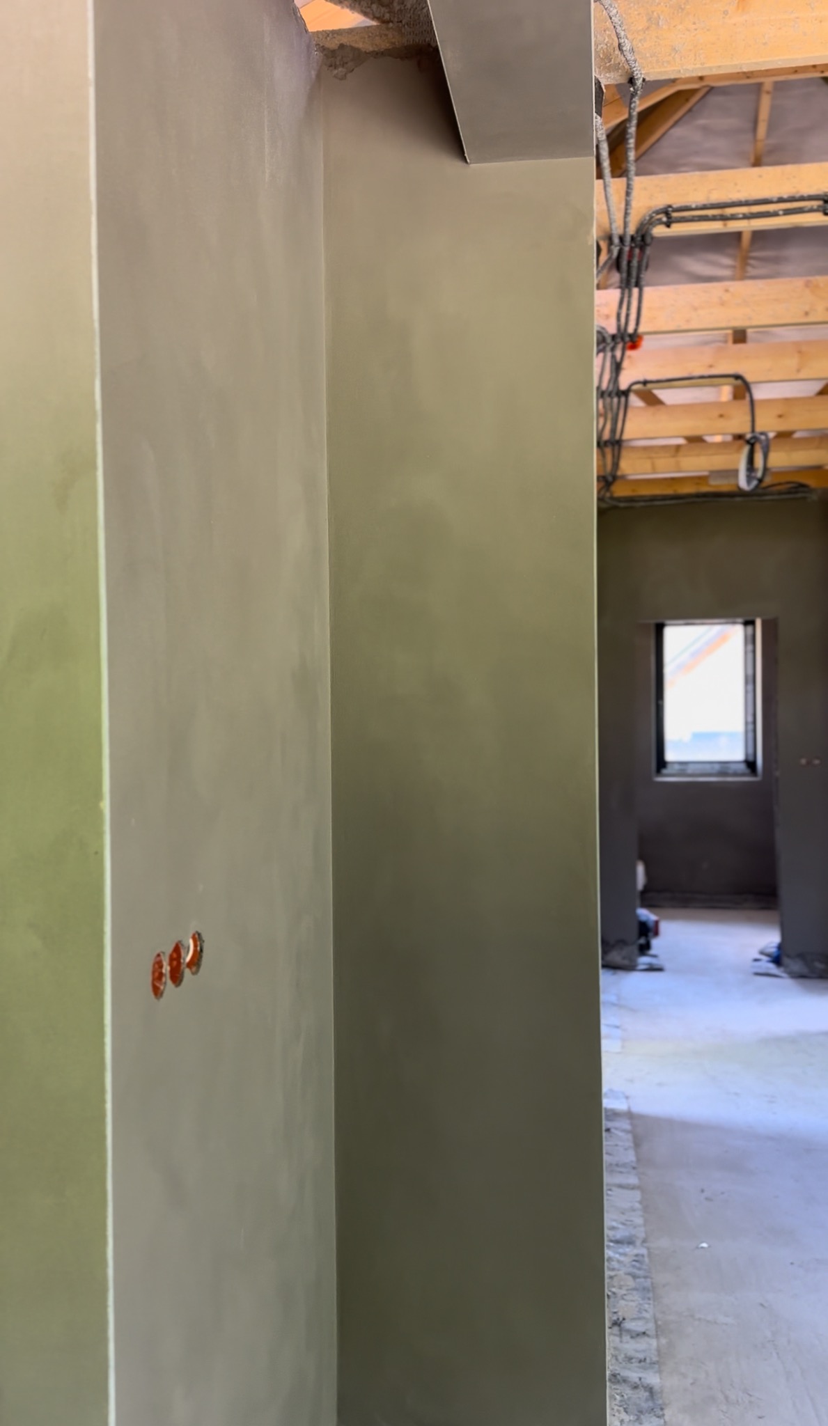 Wnętrze w trakcie prac wykończeniowych: gładkie, szare ściany z widocznymi puszkami elektrycznymi, surowy strop i okno w głębi. Minimalistyczny, surowy styl.