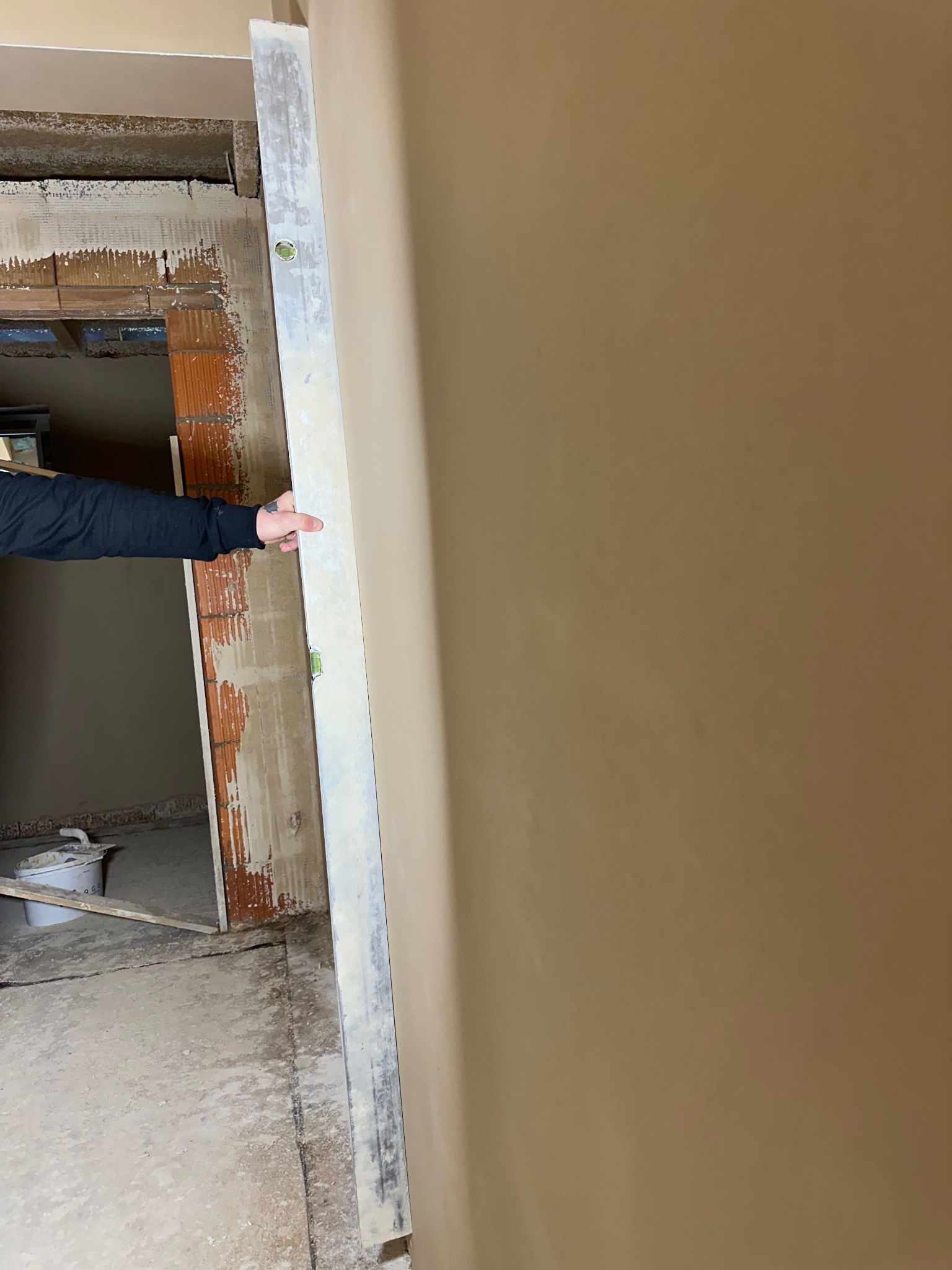 Wyrównywanie ściany gładzią przy użyciu poziomicy, widoczny fragment muru i wiadro z zaprawą. Praca w trakcie wykańczania wnętrza.