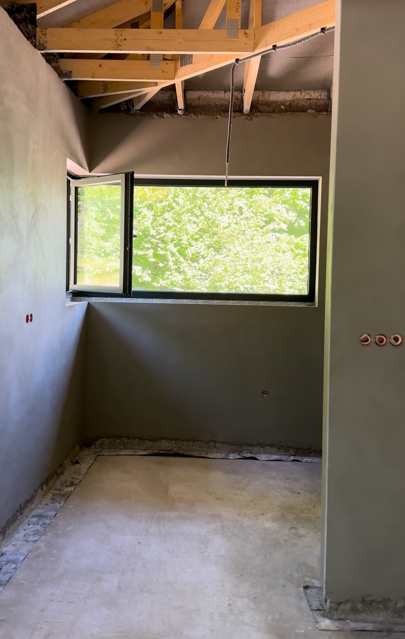 Wnętrze w trakcie tynkowania: szare ściany, otwarte okno z widokiem na zieleń, surowy sufit z drewnianą konstrukcją. Widoczne gniazdka elektryczne.