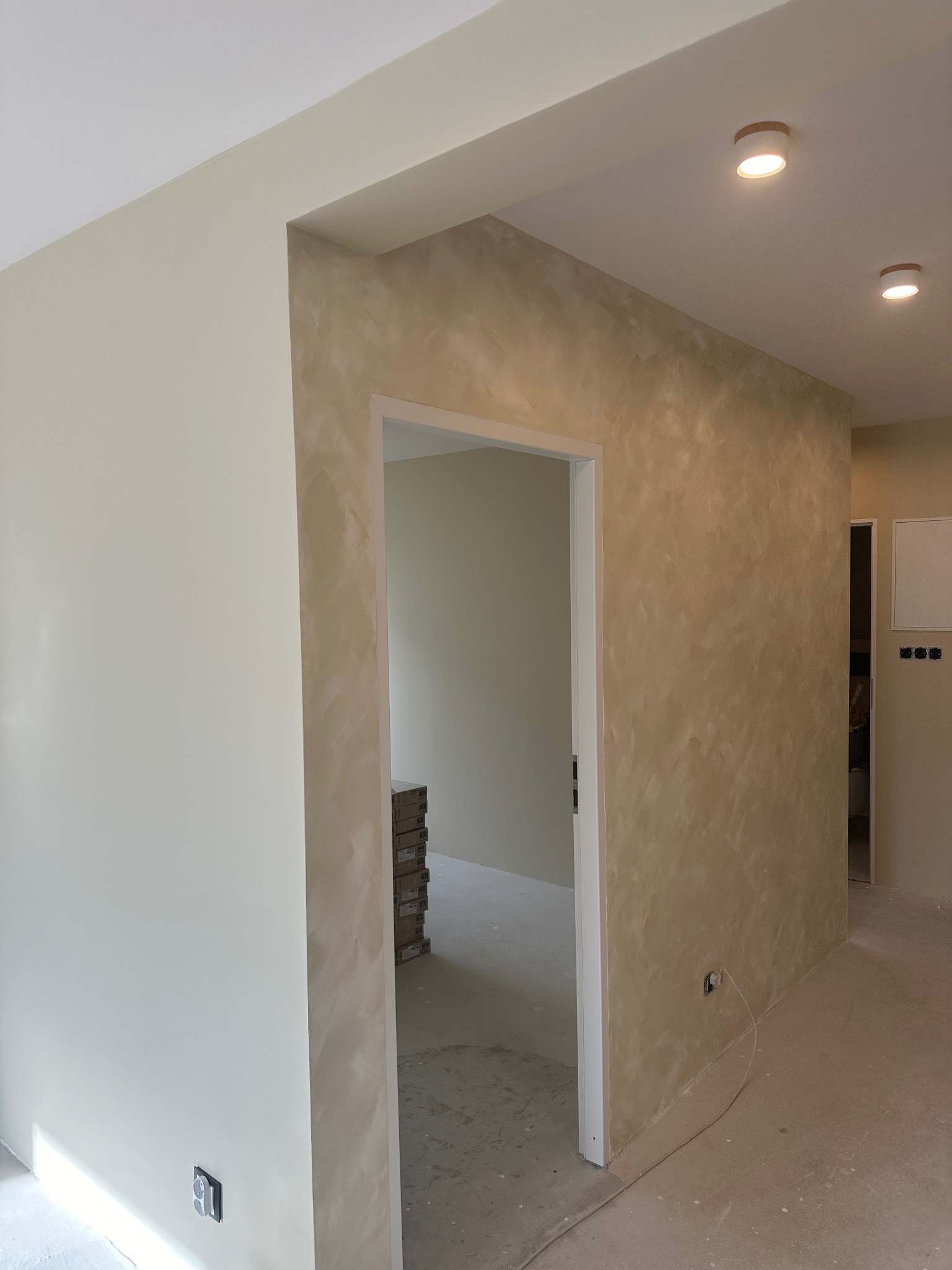Beżowa ściana z efektem dekoracyjnym w nowym wnętrzu. Widoczne gniazdka elektryczne i fragment podłogi. W tle otwarte drzwi i ułożone kartony.