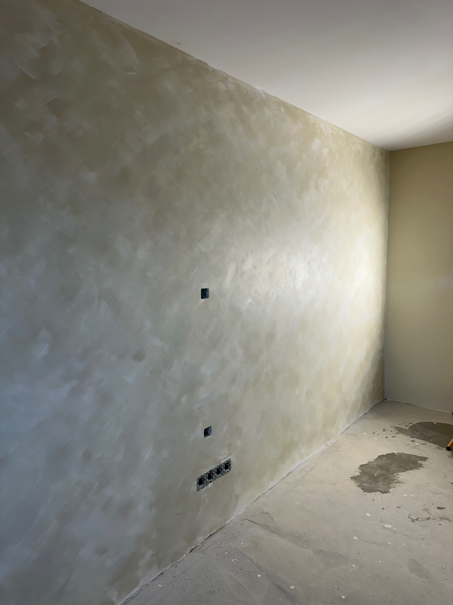 Ściana po nałożeniu dekoracyjnej gładzi w odcieniach beżu i szarości, z widocznymi gniazdkami elektrycznymi. Na podłodze widoczne ślady po pracach wykończeniowych.