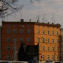 Pomarańczowy budynek mieszkalny z wieloma oknami, widziany z ulicy z samochodami, z drzewem bez liści w lewym górnym rogu i błękitnym niebem w tle.