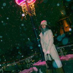 Patryk Skrzyniarz - Kobieta w płaszczu pozuje w zimowej scenerii pod neonową lampą. Śnieg pada, tworząc magiczną atmosferę. Ujęcie z dołu podkreśla wysokość lampy i sylwetkę modelki.