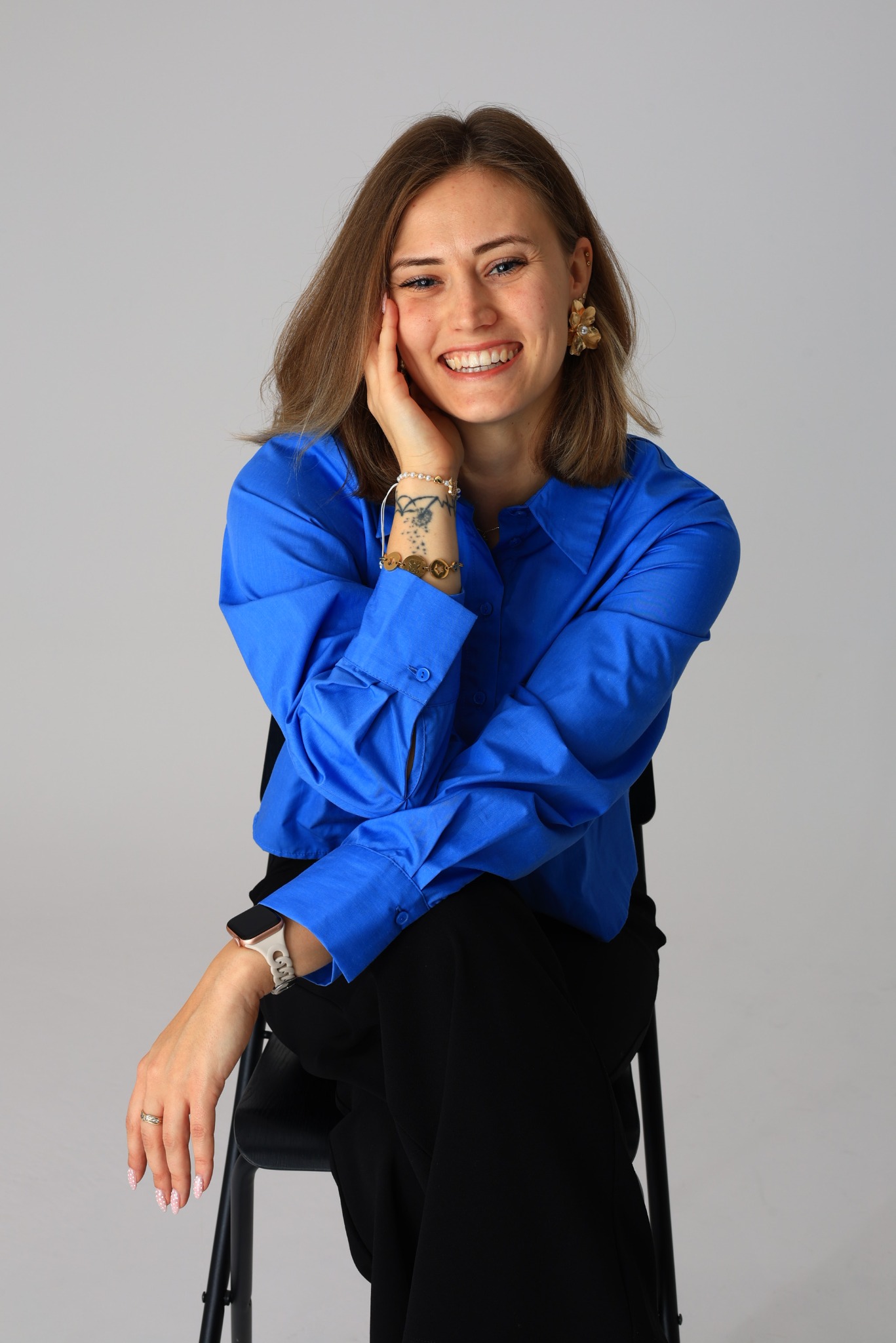 Uśmiechnięta kobieta w błękitnej koszuli siedzi na krześle, opierając twarz na dłoni. Widoczny tatuaż na przedramieniu, złote kolczyki i zegarek na ręce.