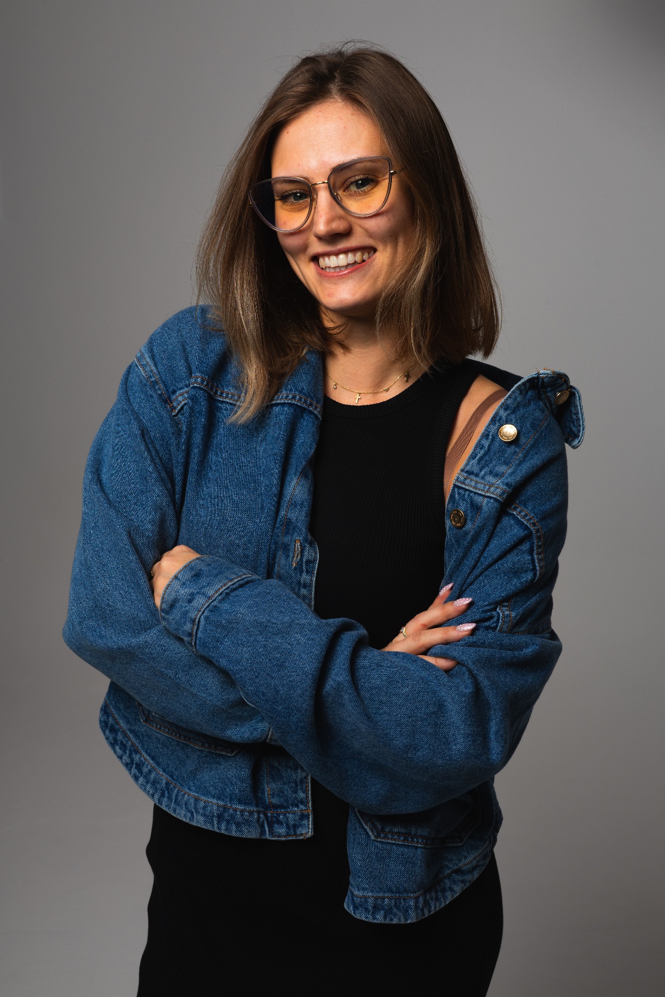Uśmiechnięta kobieta w okularach i jeansowej kurtce, pozująca na szarym tle. Widoczny złoty łańcuszek z krzyżykiem i starannie wykonany manicure.