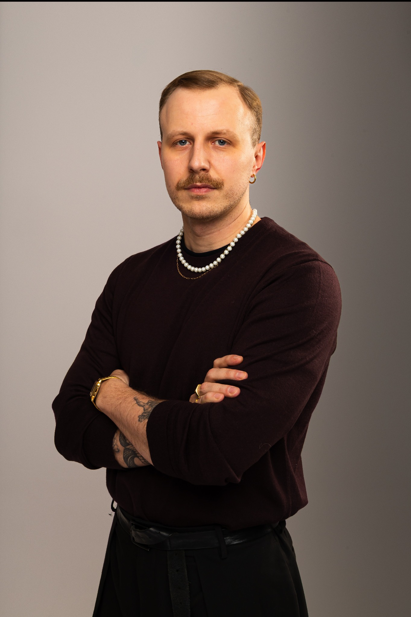 Portret mężczyzny z wąsami, w bordowym swetrze i perłach, na szarym tle. Złoty zegarek i pierścionek dodają elegancji. Sesja biznesowa.