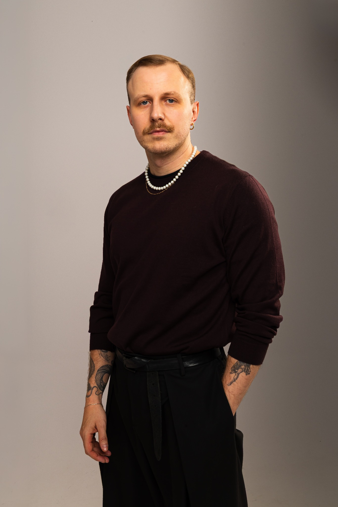 Mężczyzna z wąsami, w bordowym swetrze i czarnych spodniach, pozuje na szarym tle. Widoczne tatuaże na rękach i perłowy naszyjnik. Ujęcie portretowe, studyjne.