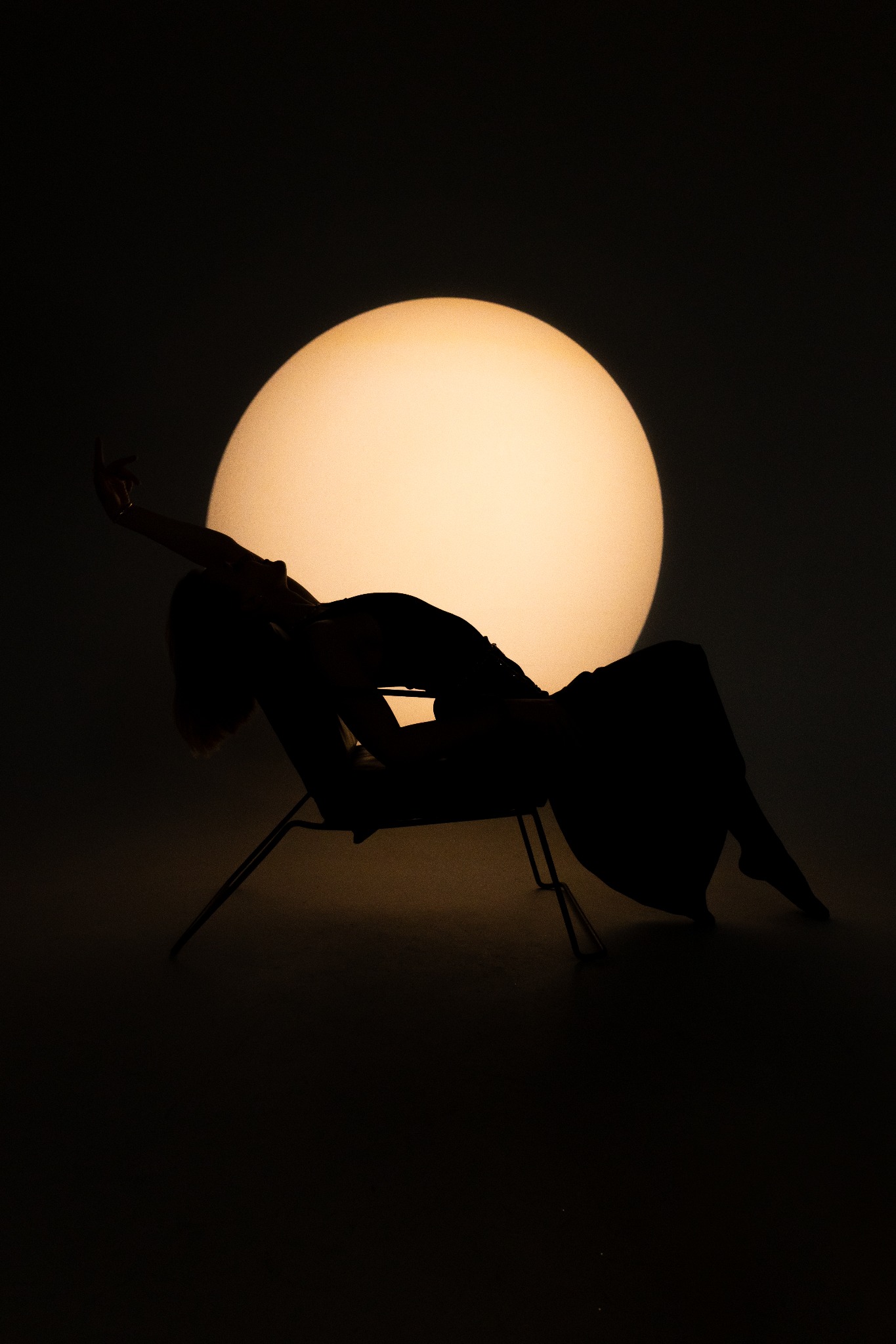 Sylwetka kobiety w fotelu na tle okrągłego światła, tworząca artystyczny portret w Warszawie. Ciemne tło podkreśla kształty i dodaje dramatyzmu.