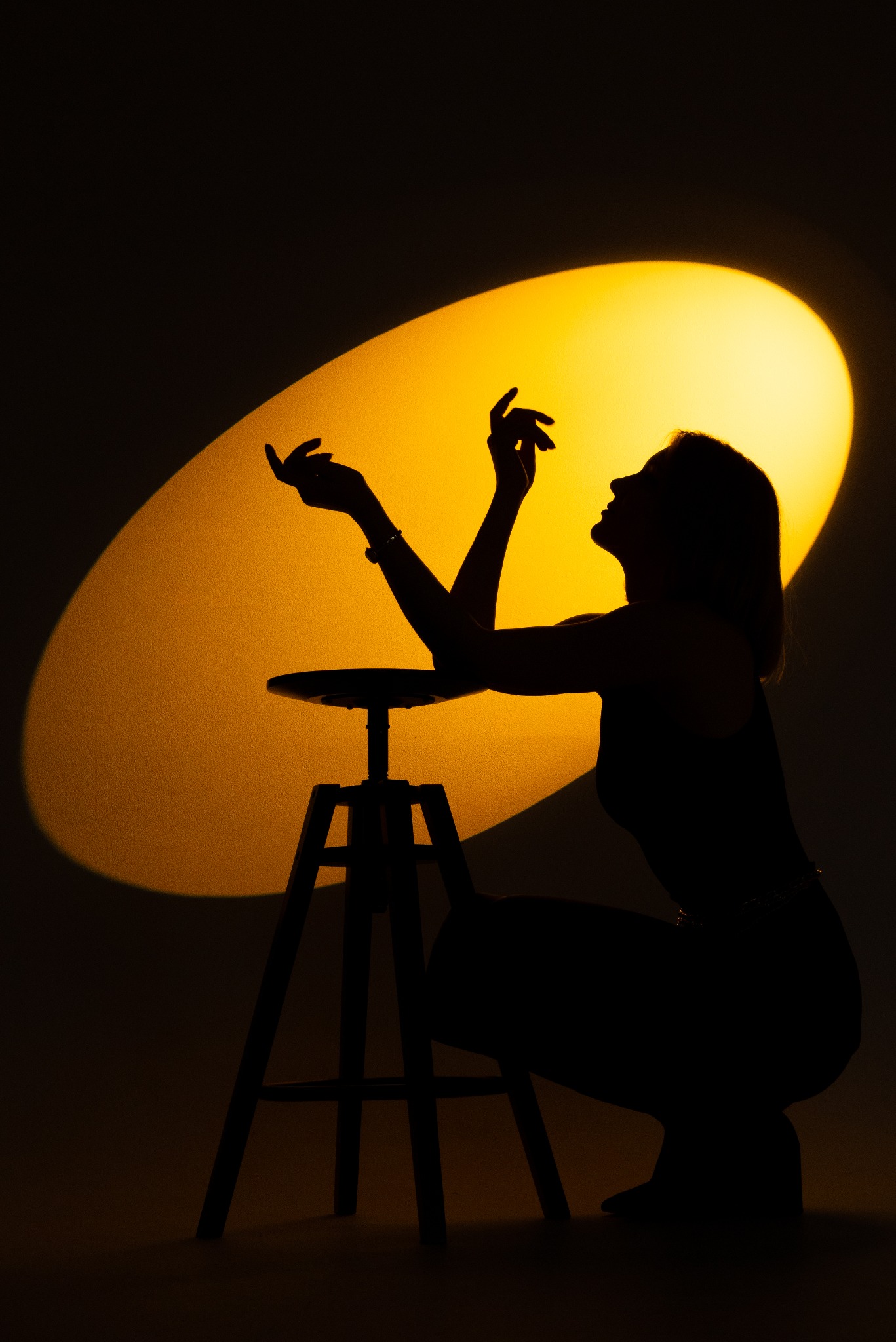 Sylwetka kobiety w kucki, oświetlona ciepłym, okrągłym światłem, z rękami uniesionymi ku górze, siedząca obok wysokiego stołka barowego. Kontrastowe oświetlenie.