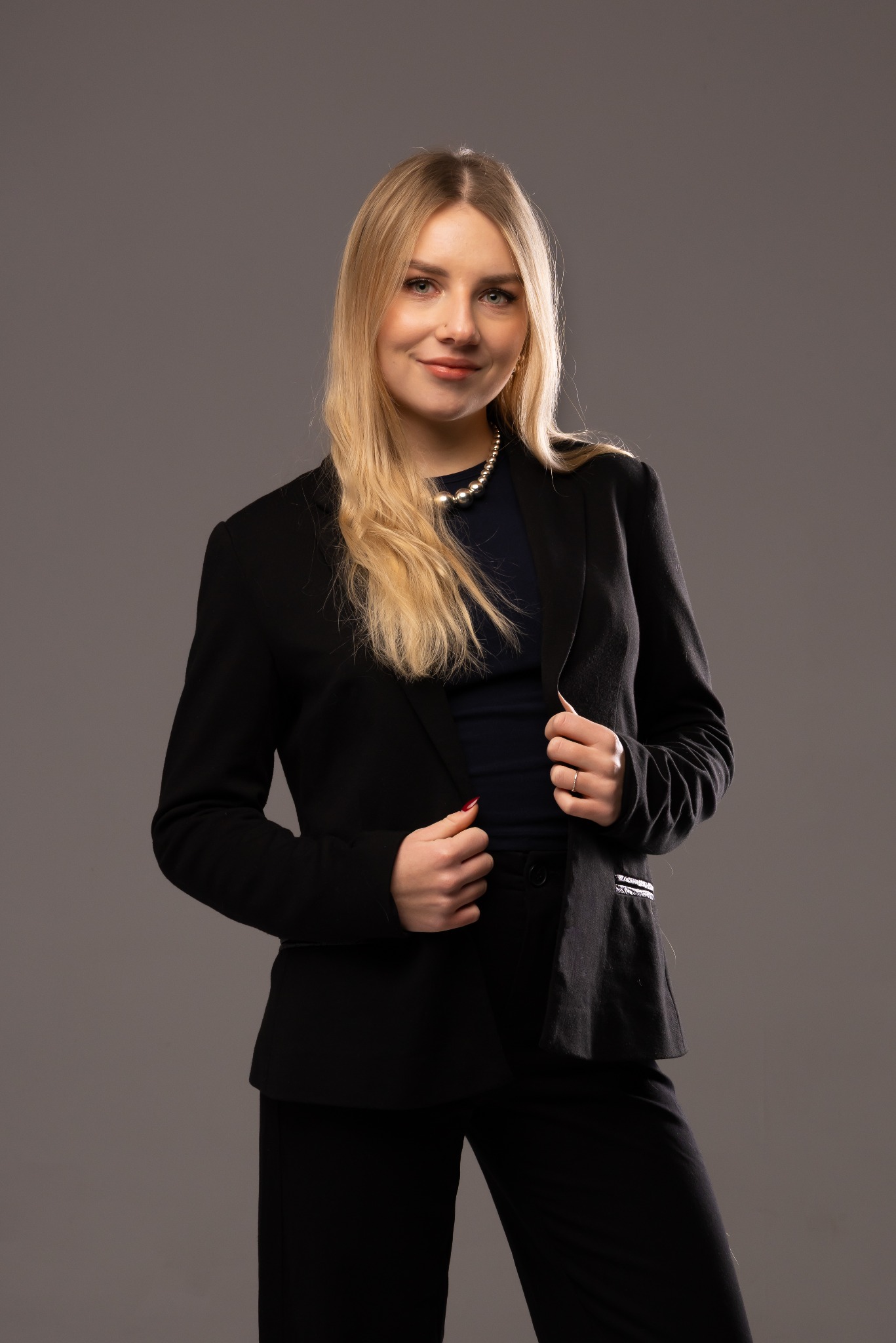Portret kobiety w czarnym garniturze i perłach na szarym tle. Uśmiechnięta blondynka z delikatnym makijażem, pozuje, trzymając marynarkę. Profesjonalny wizerunek.
