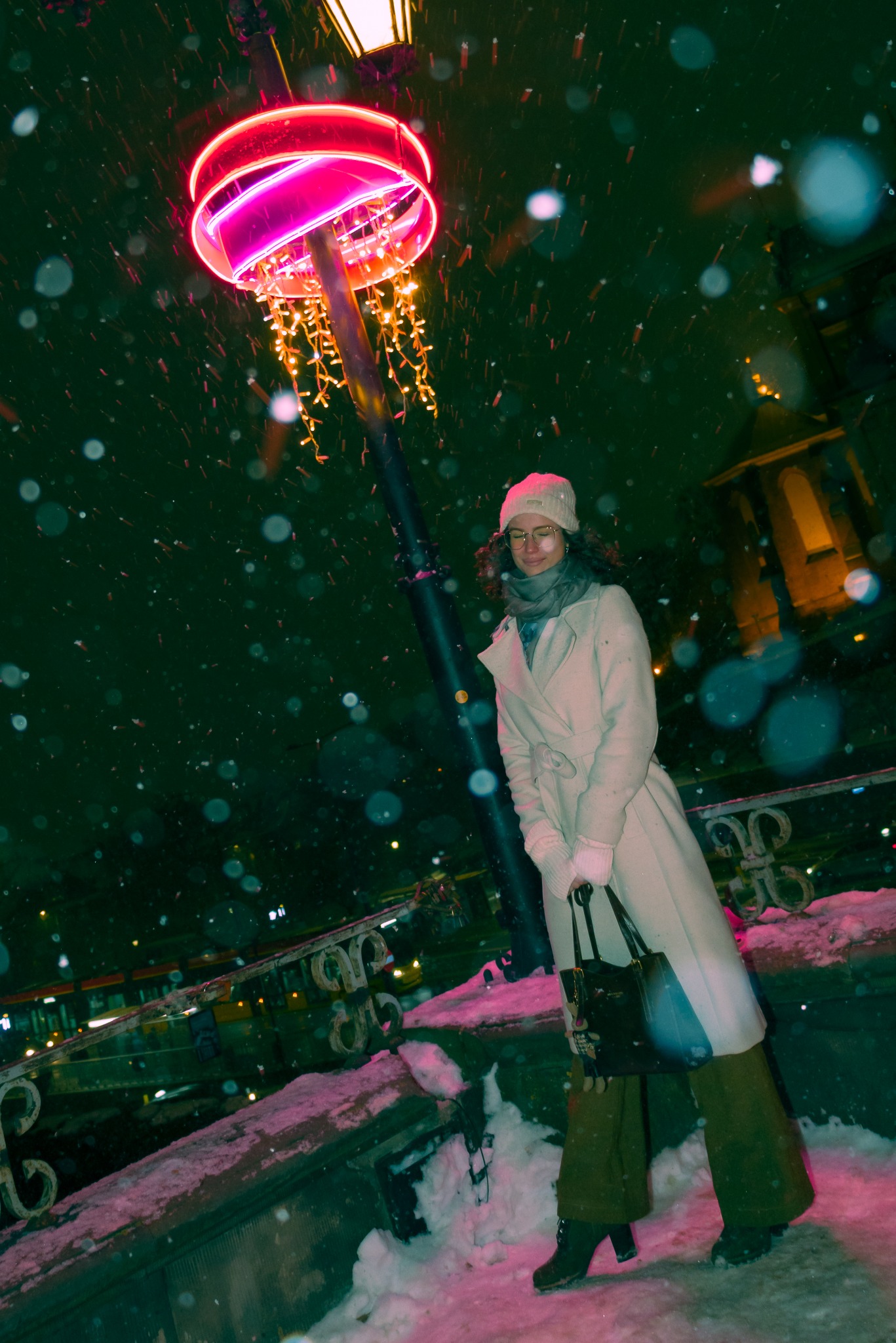 Kobieta w płaszczu pozuje w zimowej scenerii pod neonową lampą. Śnieg pada, tworząc magiczną atmosferę. Ujęcie z dołu podkreśla wysokość lampy i sylwetkę modelki.