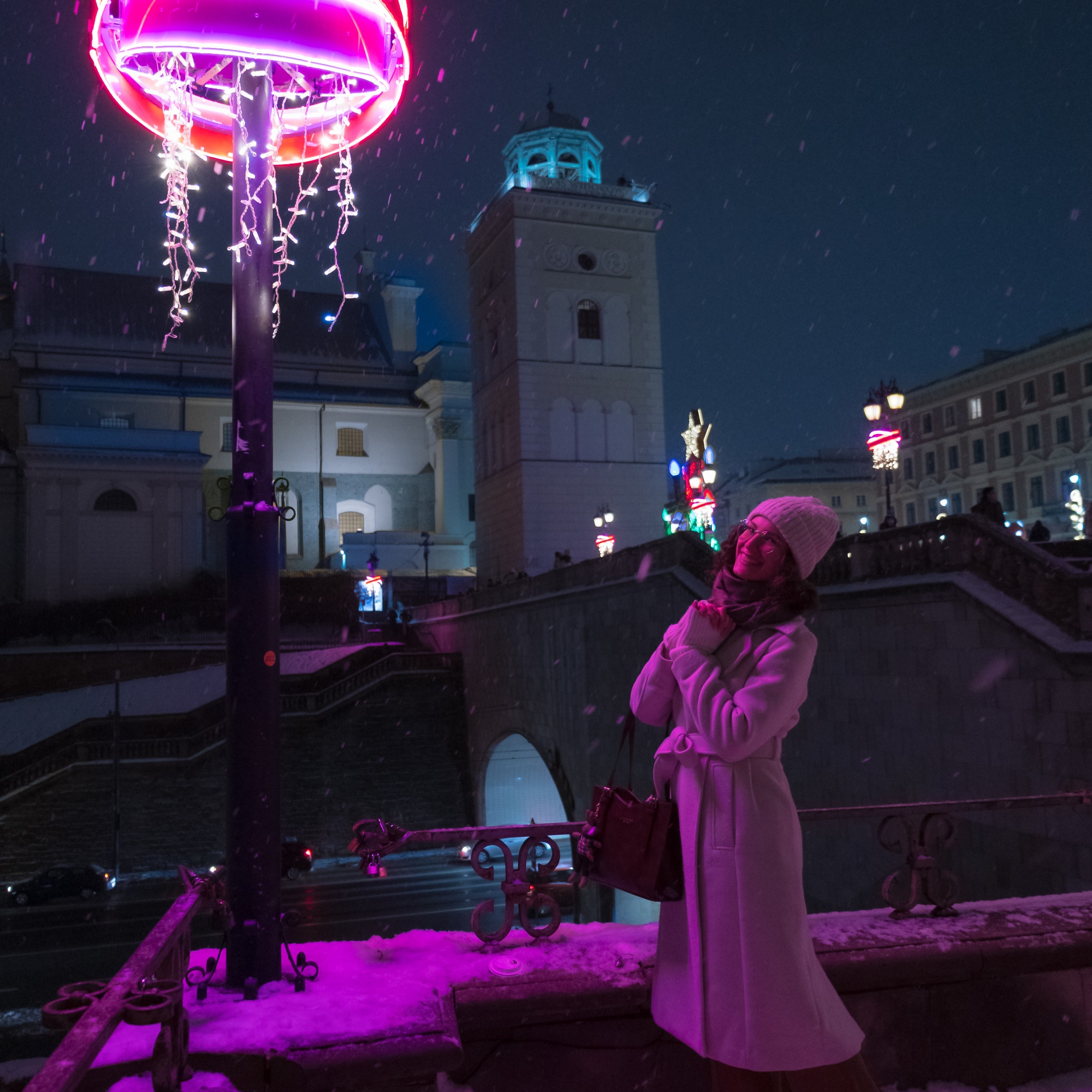 Zimowa sesja: Uśmiechnięta kobieta w białym płaszczu i czapce, podświetlona neonowym światłem na tle ośnieżonej Starówki w Warszawie podczas padającego śniegu.