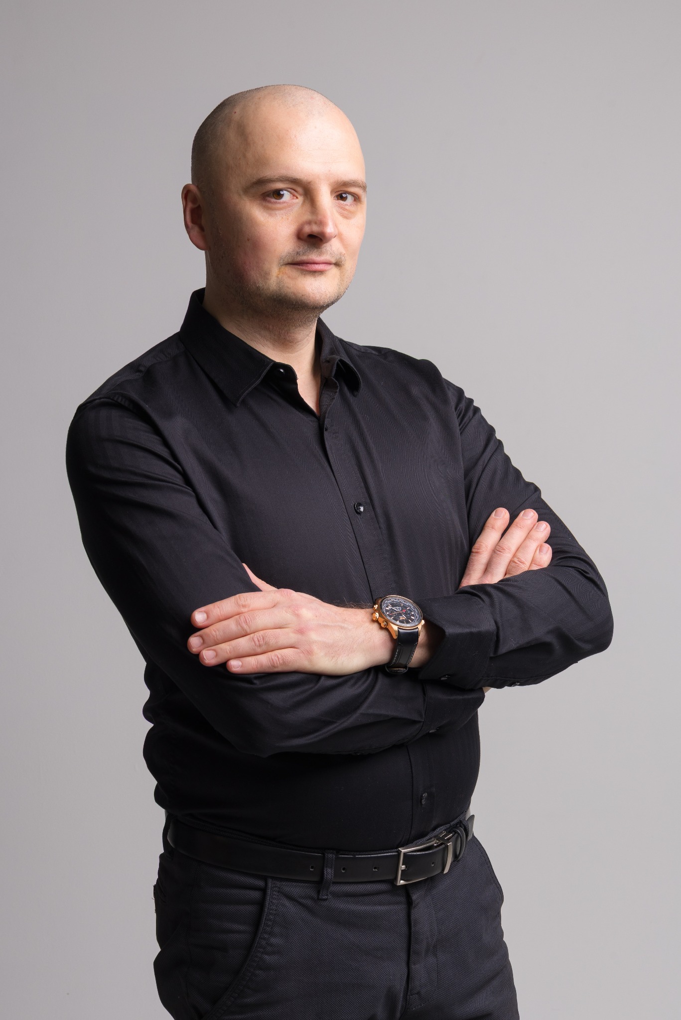 Mężczyzna w czarnej koszuli, z założonymi rękami i zegarkiem na nadgarstku, na jednolitym szarym tle. Portret biznesowy z subtelnym oświetleniem.