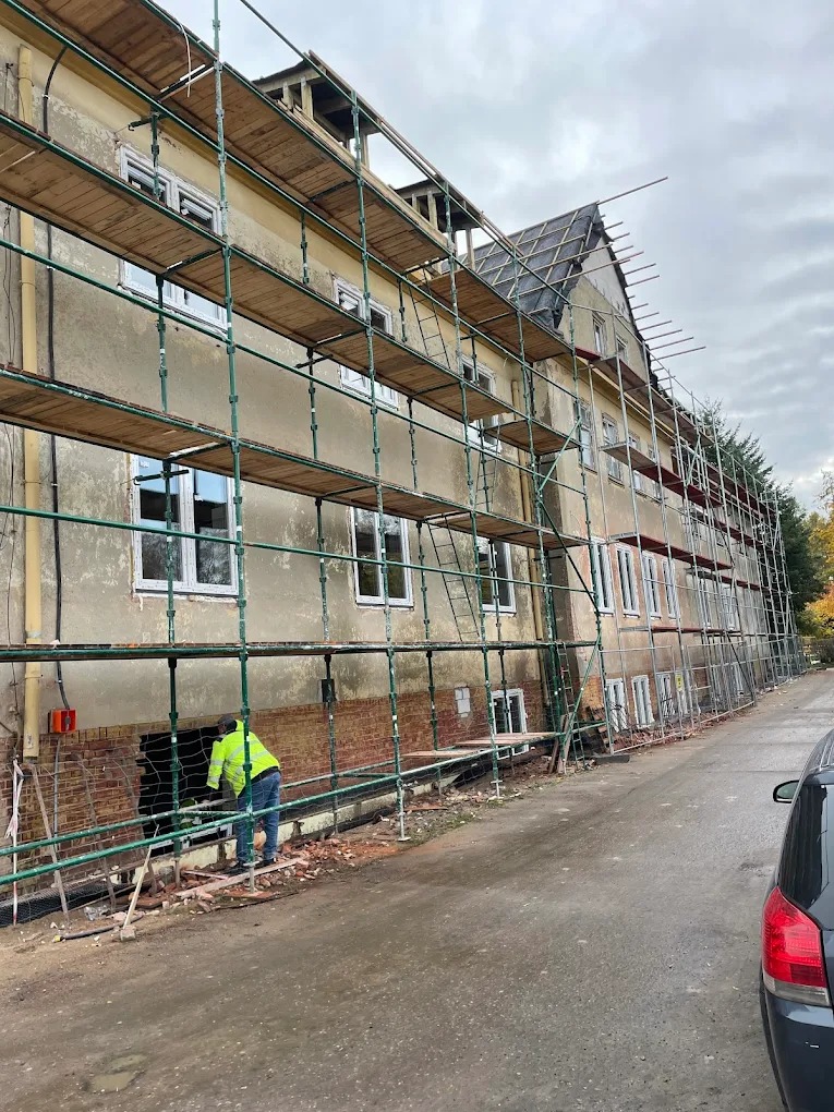 Budynek w rusztowaniach z nowymi oknami, pracownik w żółtej kamizelce przy otworze w ścianie. Widok z boku, pochmurne niebo.