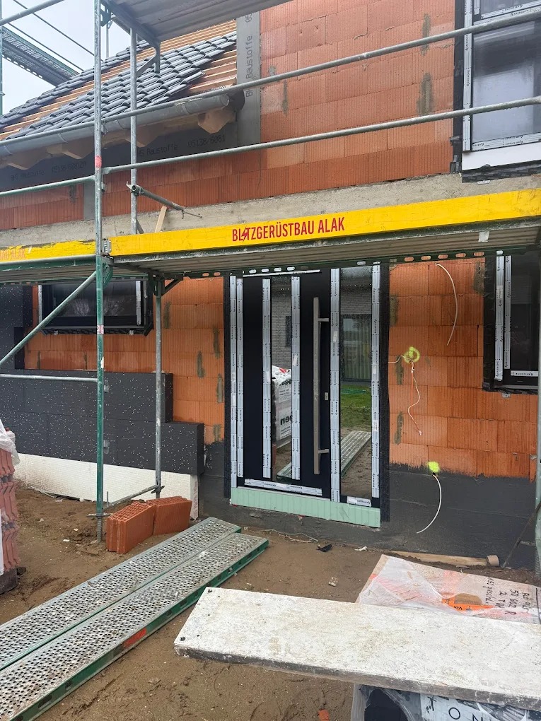 Montaż drzwi wejściowych w surowym budynku z cegły, widoczne rusztowanie i materiały budowlane. Drzwi z długim uchwytem, zabezpieczone folią.