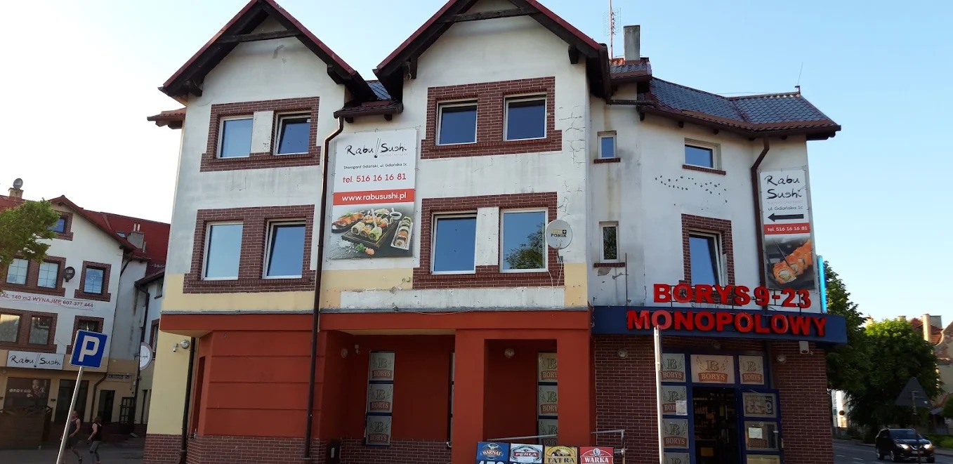 Budynek z oknami PCV w ceglanej obudowie, na którym wiszą reklamy restauracji sushi. Widoczne okna na dwóch piętrach, z jasnymi ramami i odbiciem nieba.