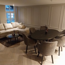 LUKABUD ŁUKASZ ŻACZEK - Eleganckie wnętrze: narożna sofa, okrągły stół z krzesłami, parkiet w jodełkę. Ściany zdobione sztukaterią. Za oknem widoczne rusztowania.