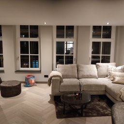 LUKABUD ŁUKASZ ŻACZEK - Jasne wnętrze salonu z parkietem w jodełkę, beżową sofą narożną, stolikiem kawowym i trzema oknami. Neutralna kolorystyka ścian i mebli.