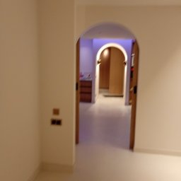 LUKABUD ŁUKASZ ŻACZEK - Białe wnętrze z łukowym przejściem do kolejnego pomieszczenia z drewnianą komodą i oświetleniem. Widoczne gniazdka elektryczne na ścianie.