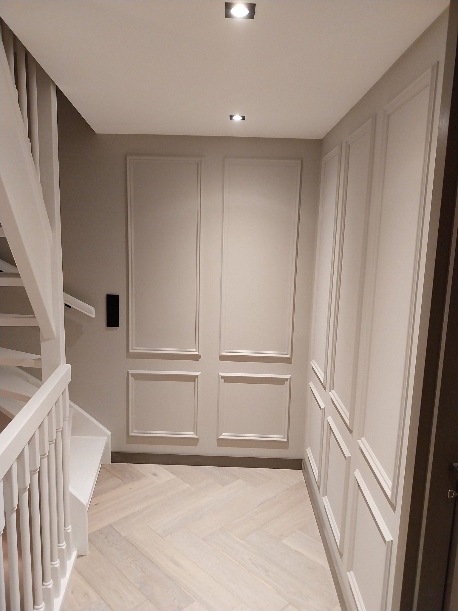 Korytarz z panelami ściennymi w stylu klasycznym, jasna podłoga ułożona w jodełkę, białe schody z balustradą. Dyskretne oświetlenie punktowe.