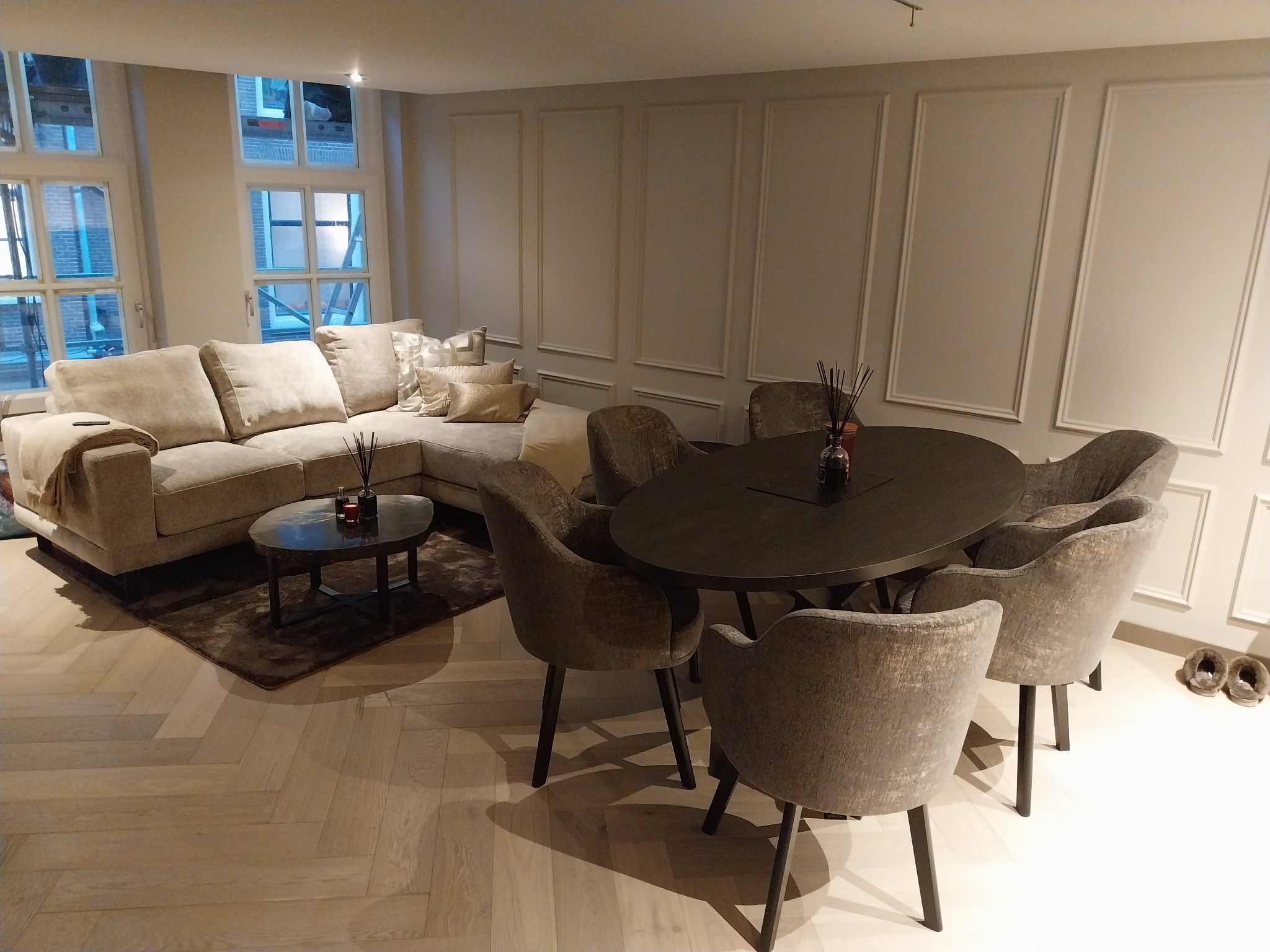 Eleganckie wnętrze: narożna sofa, okrągły stół z krzesłami, parkiet w jodełkę. Ściany zdobione sztukaterią. Za oknem widoczne rusztowania.