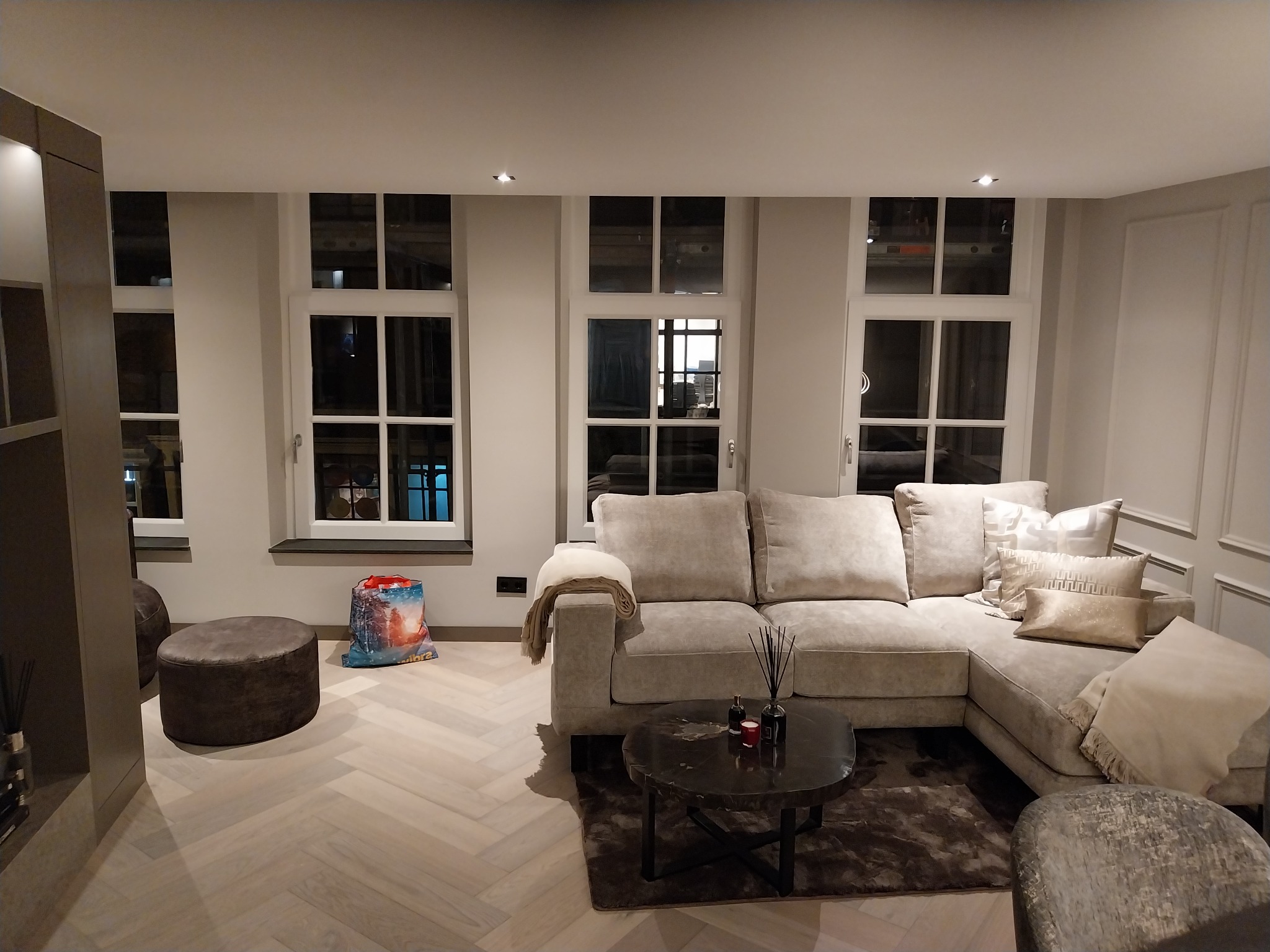 Jasne wnętrze salonu z parkietem w jodełkę, beżową sofą narożną, stolikiem kawowym i trzema oknami. Neutralna kolorystyka ścian i mebli.