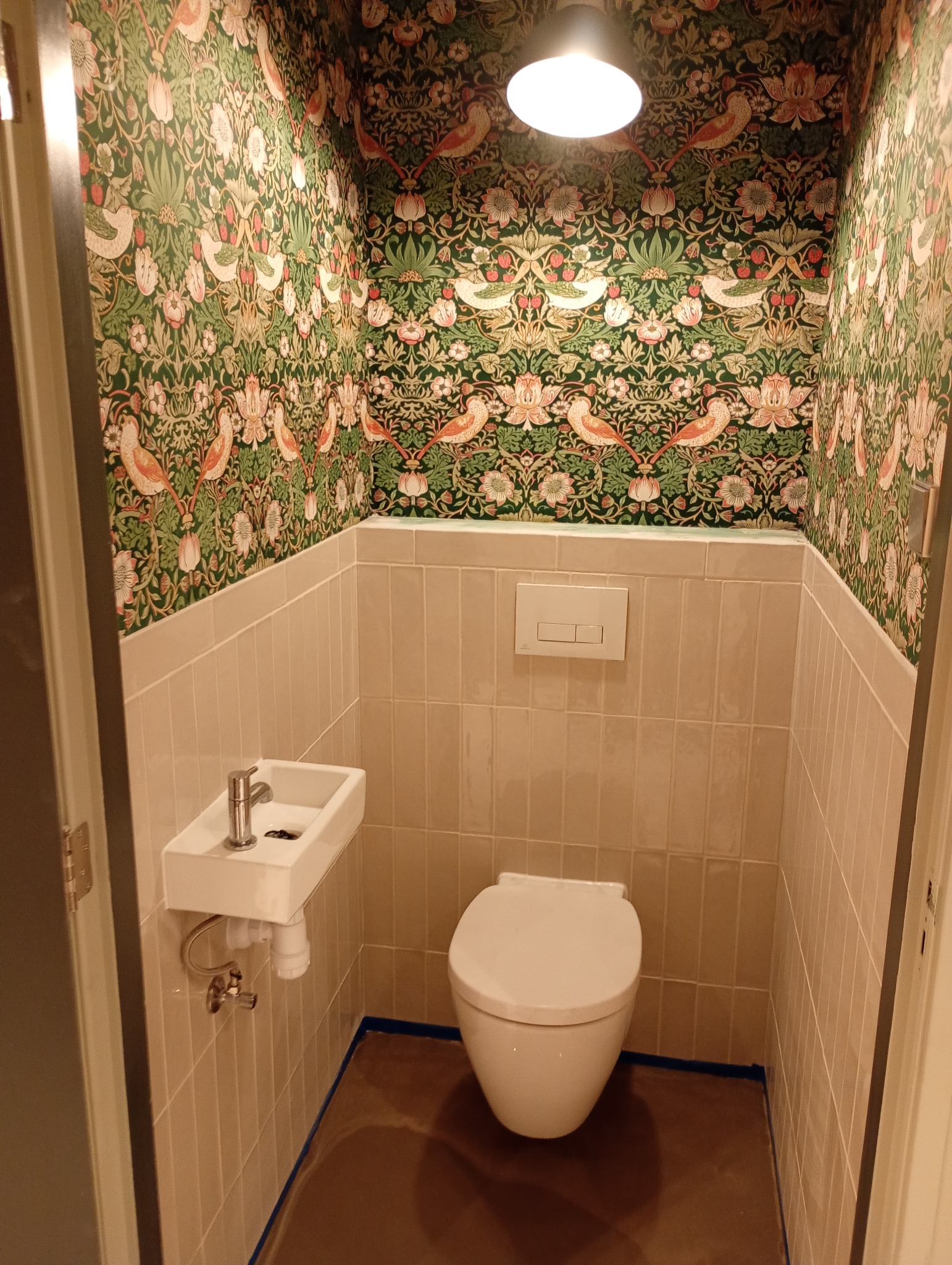 Kompaktowa toaleta z umywalką, wykończona beżowymi płytkami i tapetą w roślinno-ptasie wzory. Widoczne oświetlenie sufitowe i brązowa podłoga. Całość w małym pomieszczeniu.