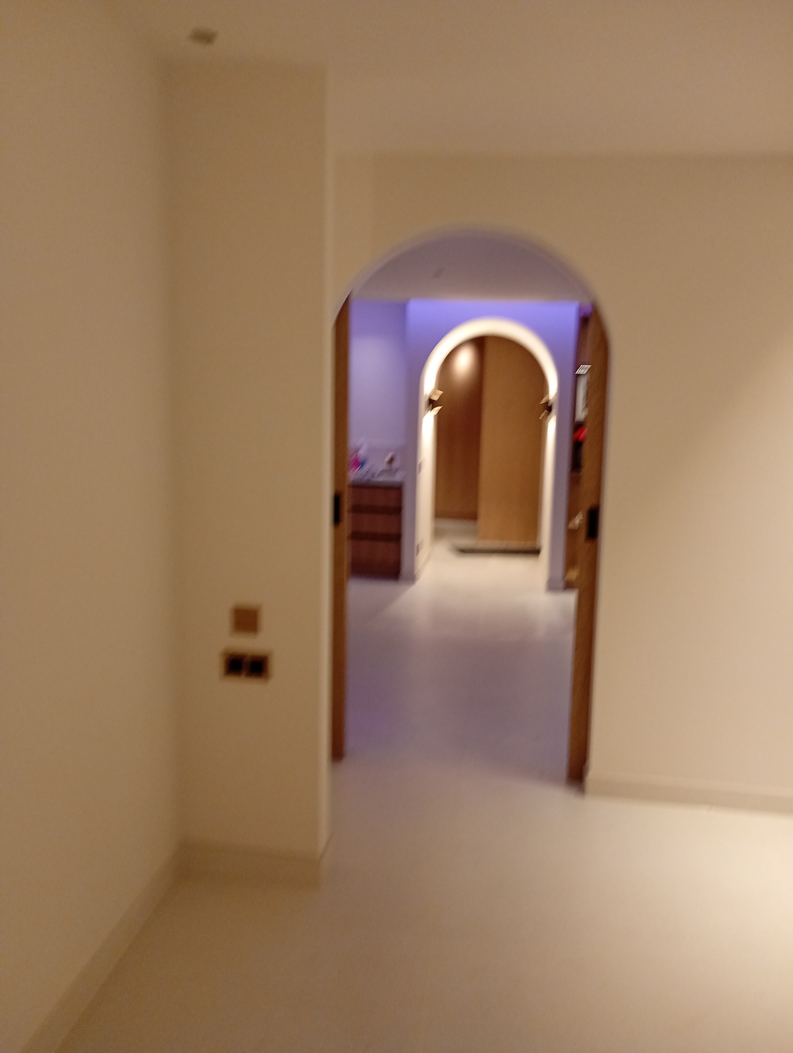 Białe wnętrze z łukowym przejściem do kolejnego pomieszczenia z drewnianą komodą i oświetleniem. Widoczne gniazdka elektryczne na ścianie.